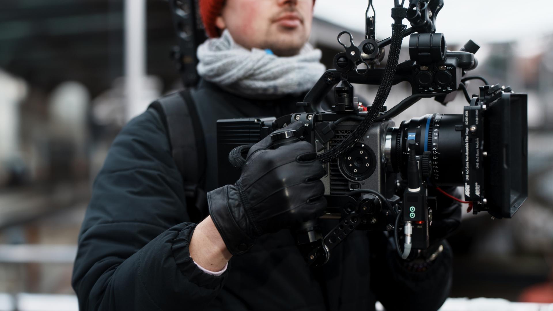 A camera operator wearing winter clothes holds a professional cinema camera rig equipped with a ZEISS lens.