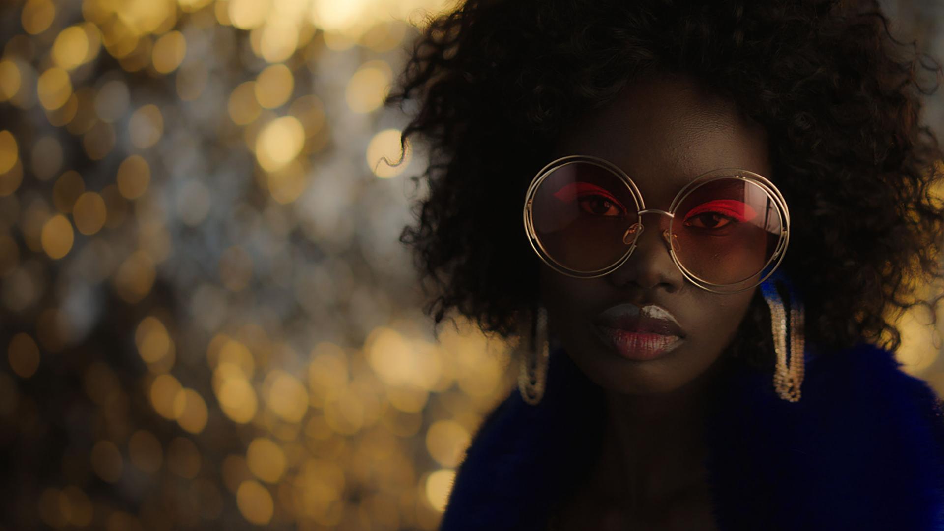 A woman wearing large, round sunglasses with red-tinted lenses and statement earrings poses in front of a golden, bokeh-lit background.