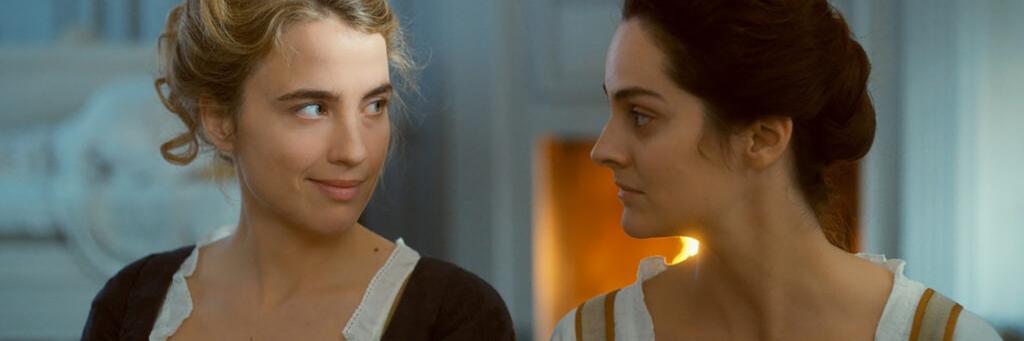 Two women in historical clothing sit indoors, warmly lit by a fireplace in the background, looking at each other.