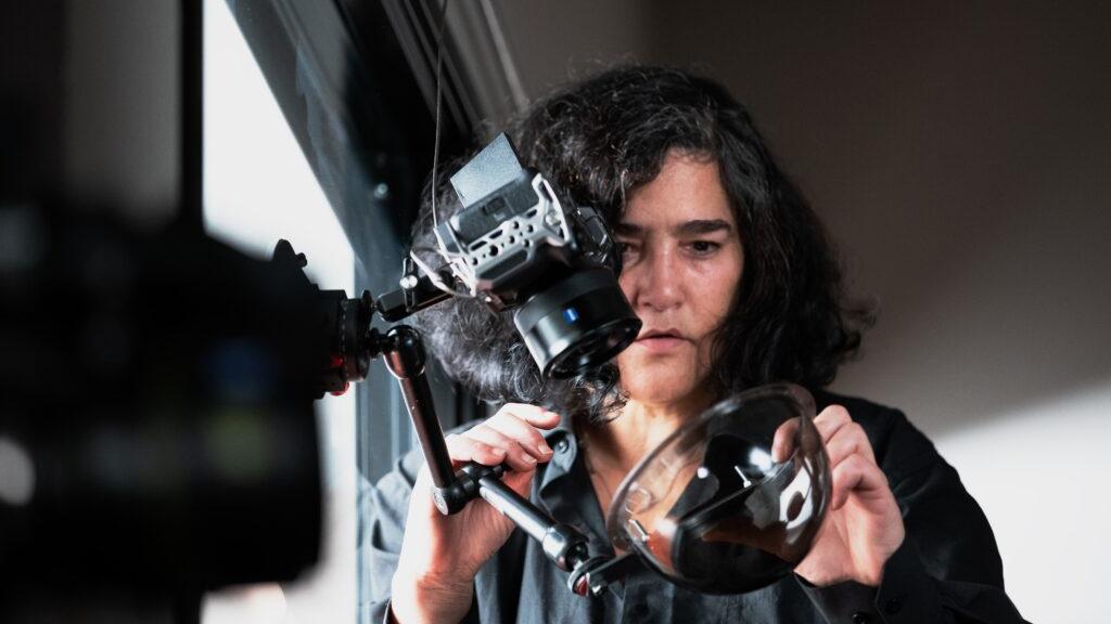 A woman is carefully adjusting a camera rig with a ZEISS lens attached, holding a large optical glass element.