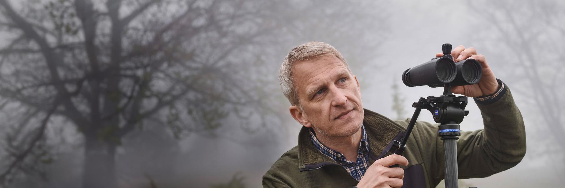A man adjusts ZEISS binoculars mounted on a tripod in a foggy, wooded outdoor setting.