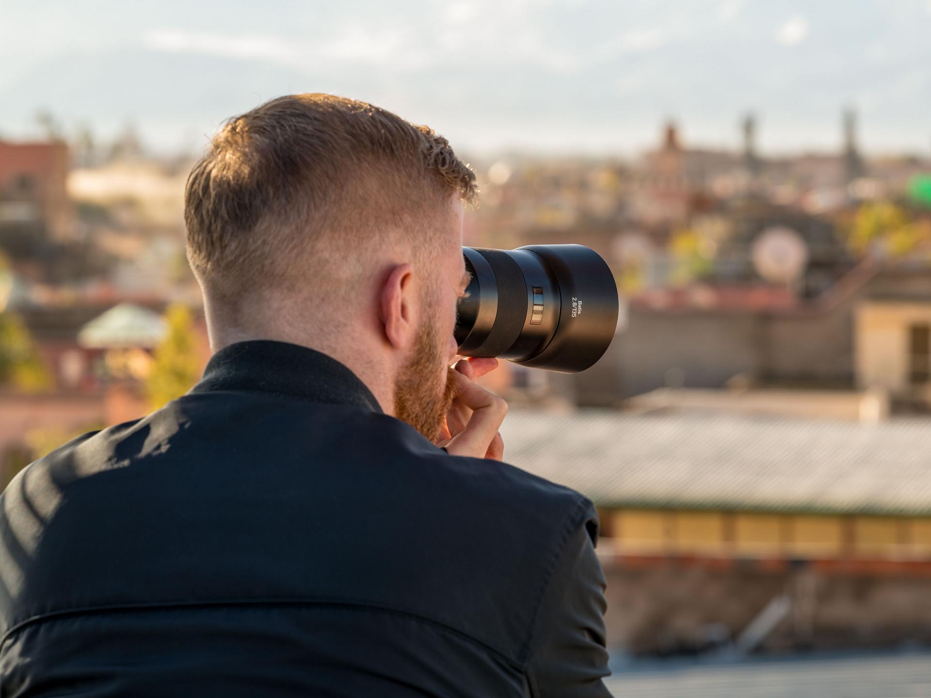 Man using a ZEISS camera lens to photograph a cityscape from an elevated outdoor location.