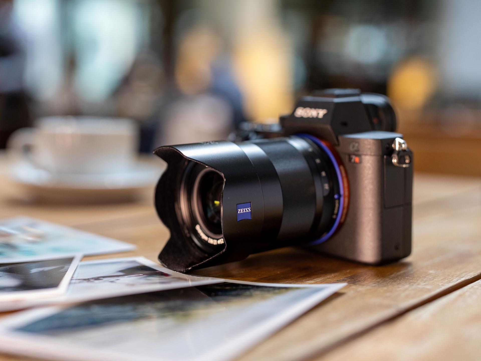 A close-up of a SONY mirrorless camera with a ZEISS lens placed on a wooden table next to printed photographs and a coffee cup in the background.