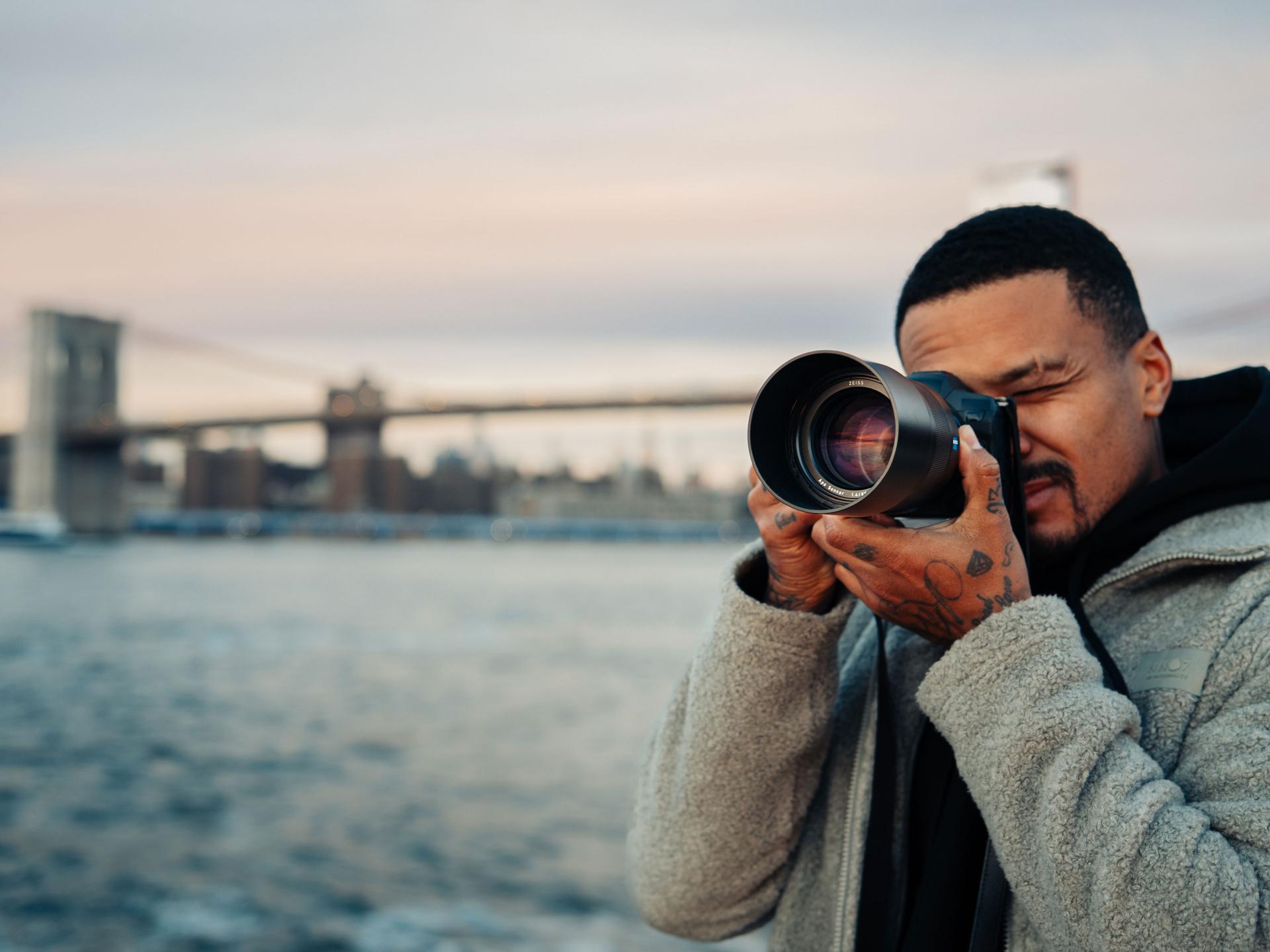 A man takes a photo with a professional camera equipped with a ZEISS lens near a waterfront with a bridge in the background.