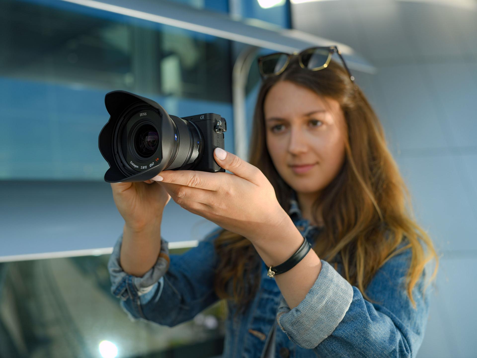 A woman holding a camera equipped with a ZEISS lens, preparing to take a photo.