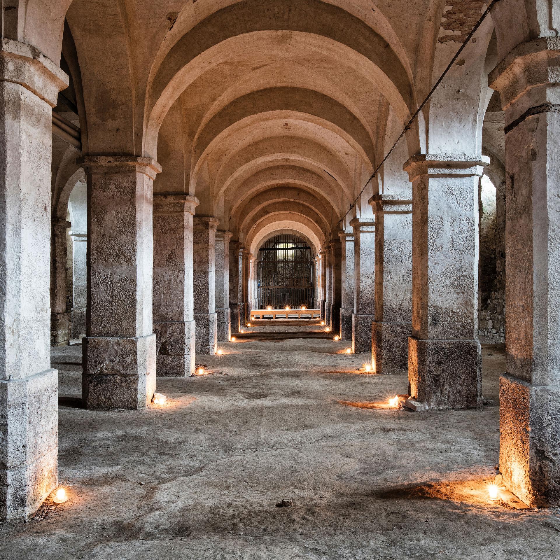 Stone corridor with a series of arches and columns, illuminated by soft ground lighting.