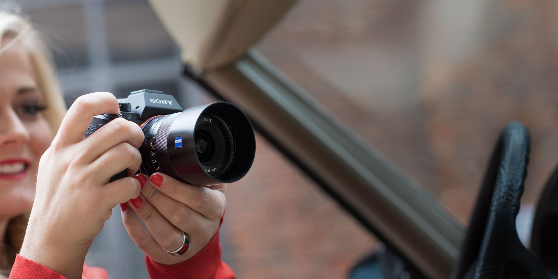 A woman is holding a SONY camera equipped with a ZEISS lens, aiming it through a car window.