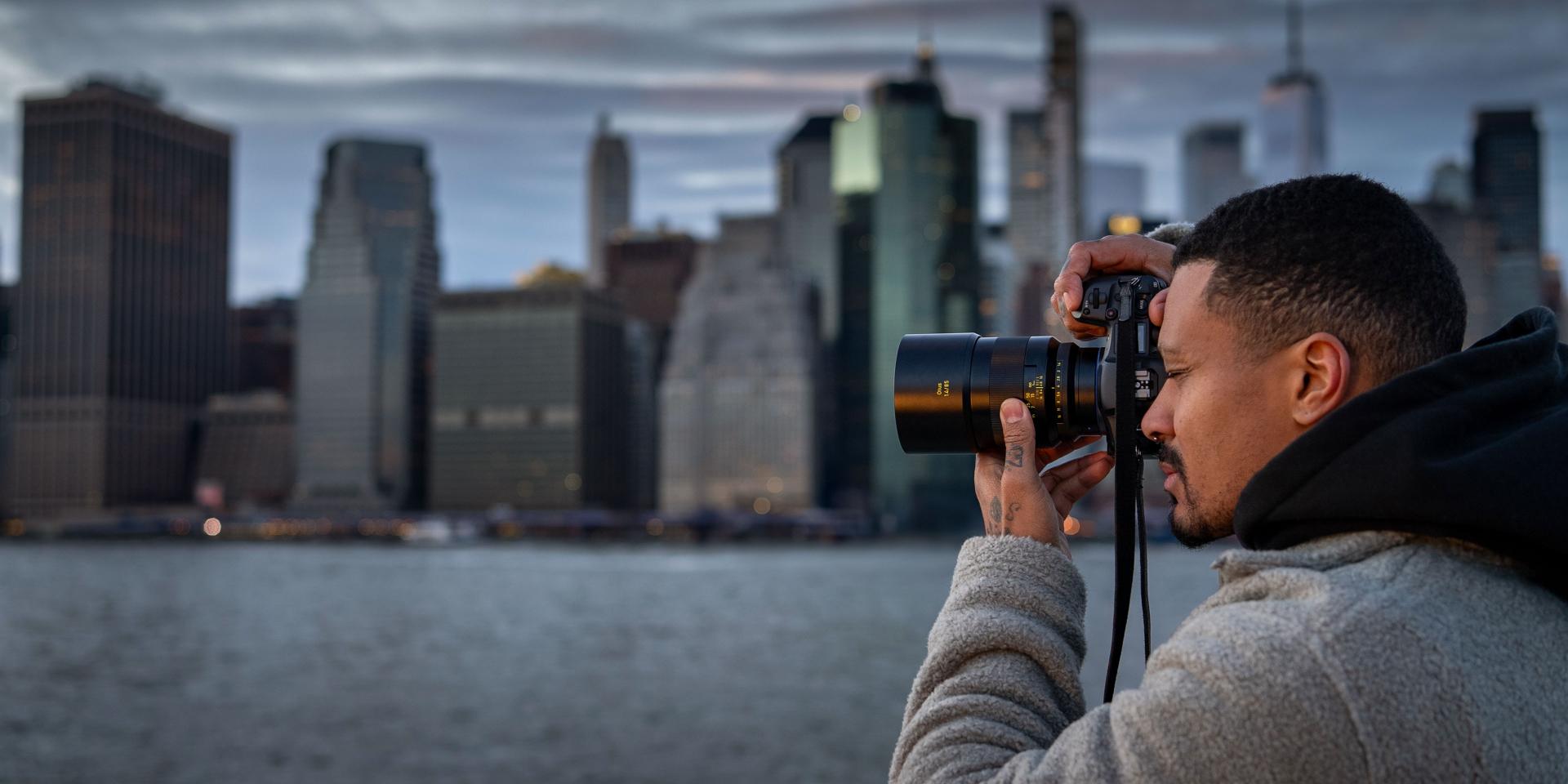A man is photographing a city skyline across the water using a camera equipped with a ZEISS lens.