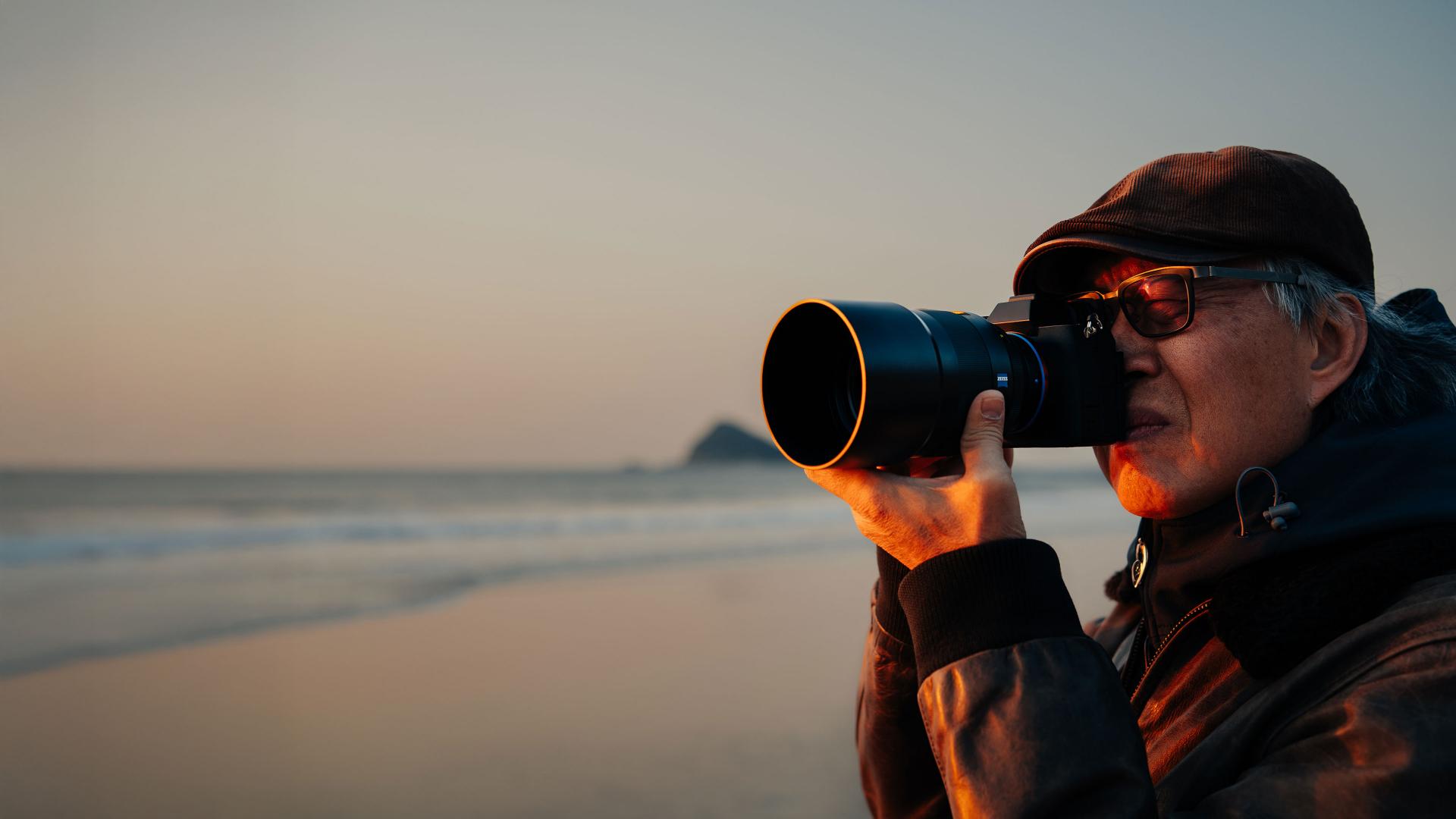 An older man wearing glasses and a cap is taking a photo with a camera equipped with a large ZEISS lens at the beach during sunset.