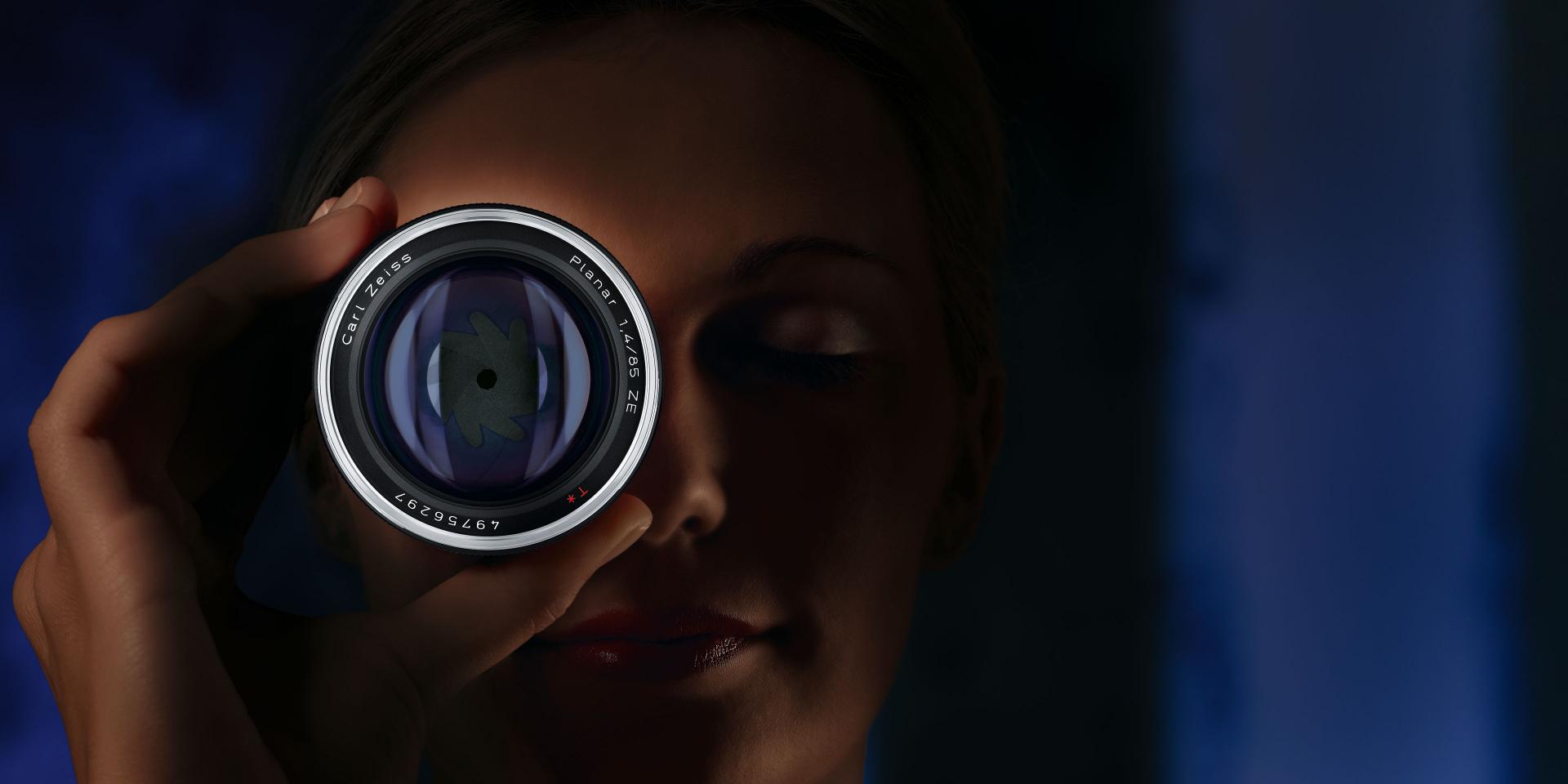 A person holding a ZEISS Planar T* 1.4/85 ZE camera lens in front of one eye.