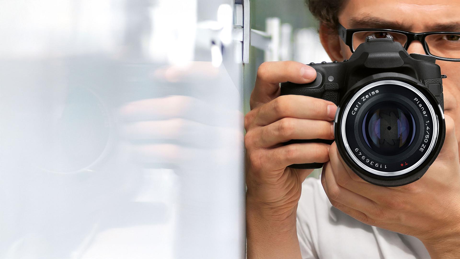 A person holding a professional DSLR camera equipped with a ZEISS lens, aiming it toward a reflective surface.