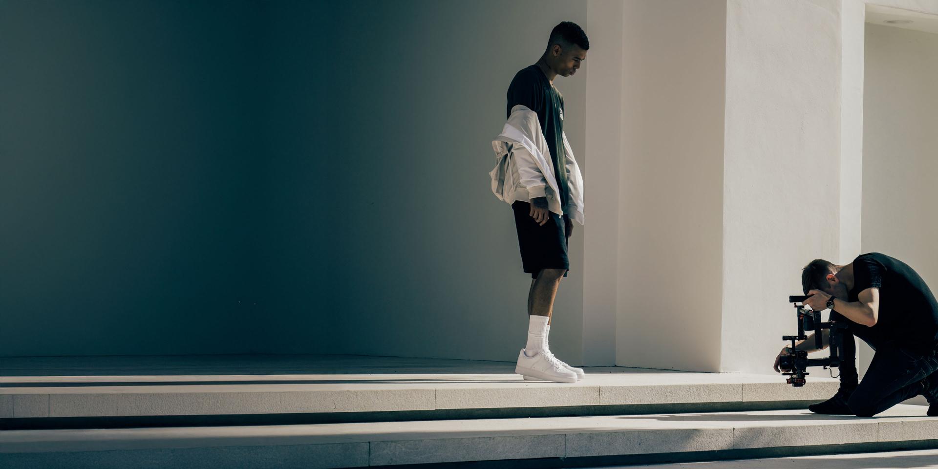 A videographer kneels on outdoor steps filming a person standing in white sneakers using a professional camera rig, with strong sunlight casting shadows.