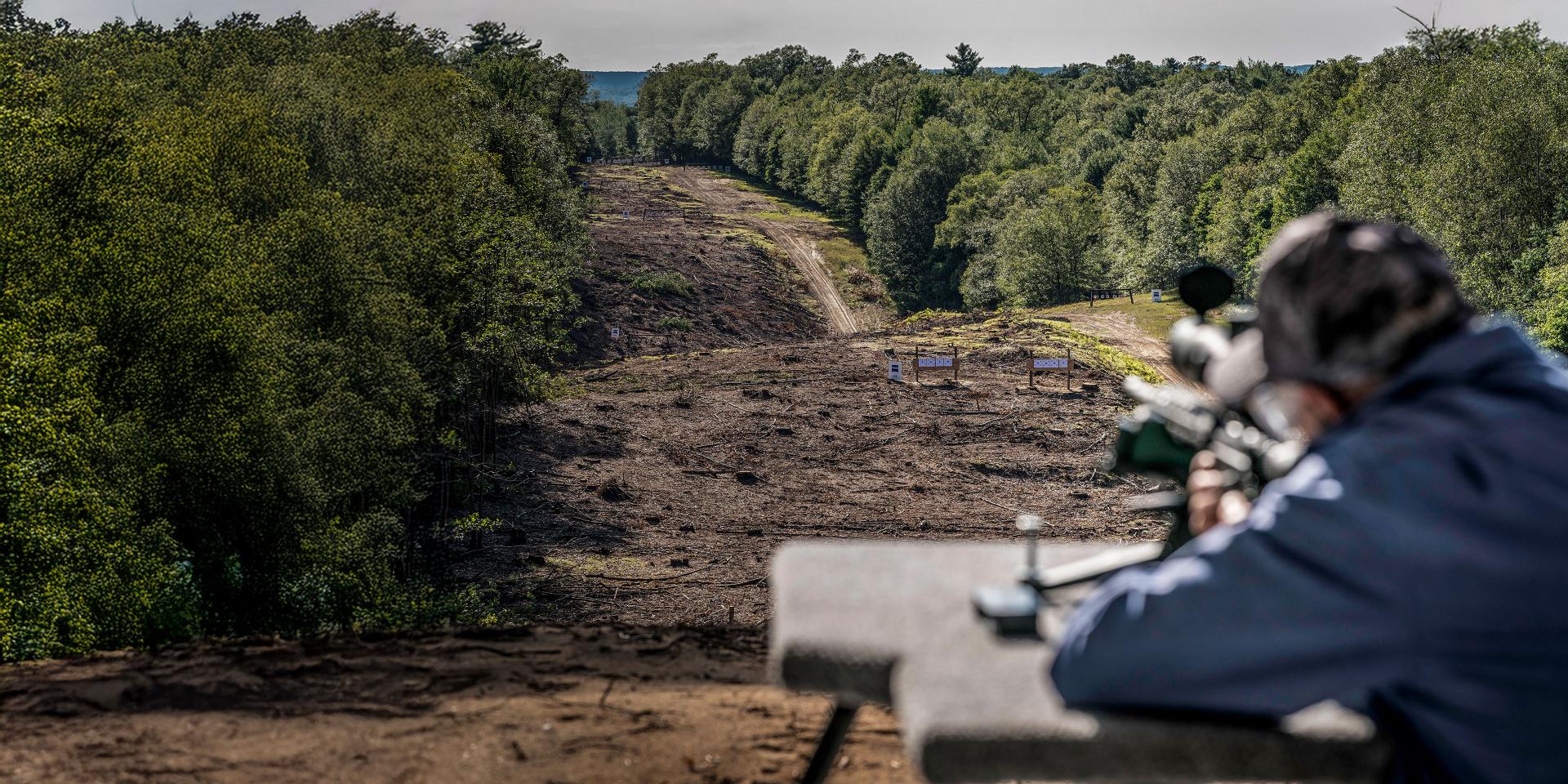 A blurred ZEISS riflescope is aimed at distant targets set in a grassy, hilly landscape under a partly cloudy sky.