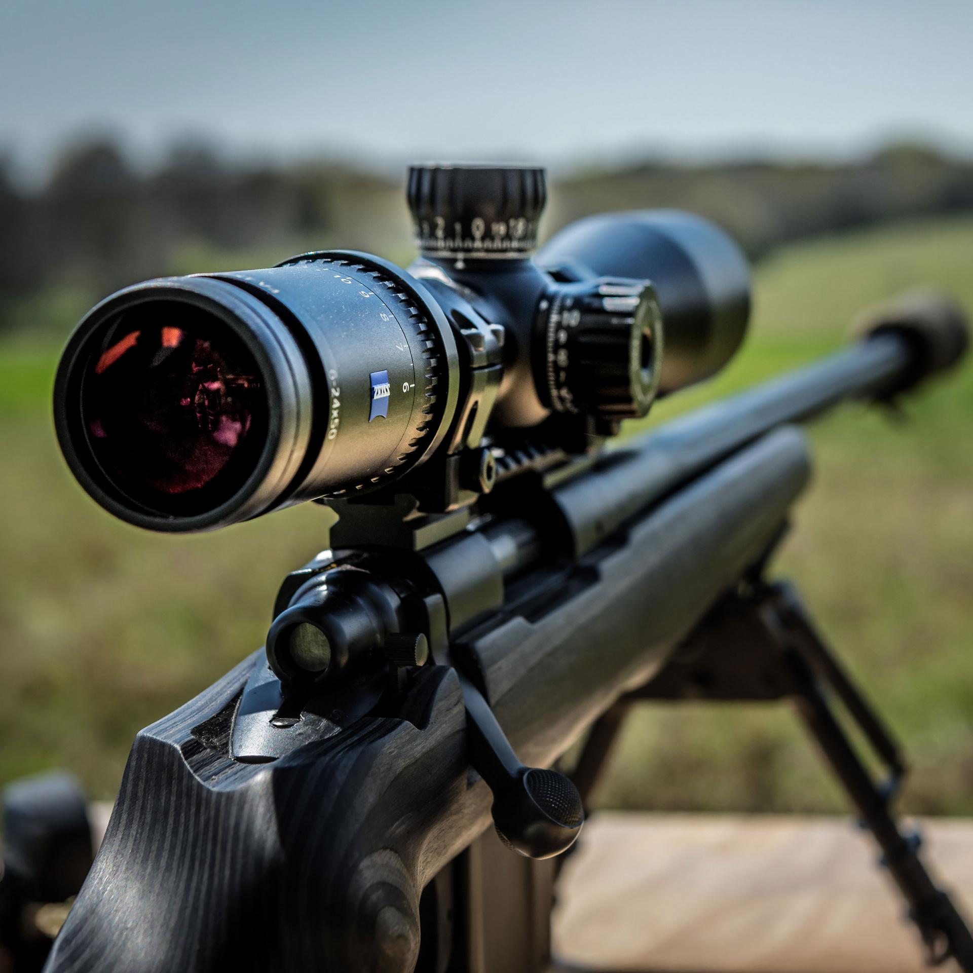 Close-up of a precision rifle equipped with a ZEISS riflescope mounted on a bipod, set against a blurred outdoor landscape.