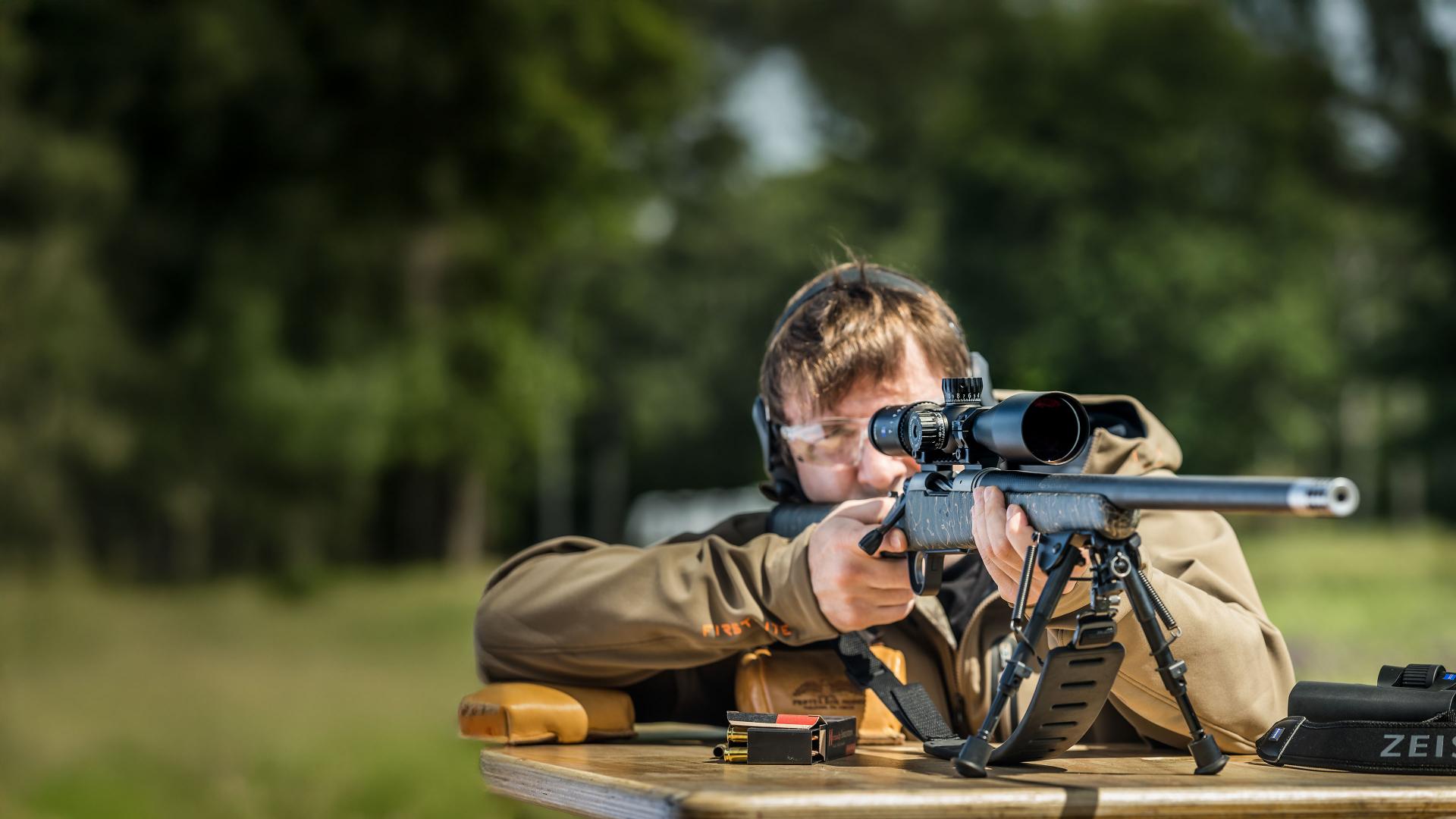 A person aims a rifle equipped with a ZEISS riflescope while wearing protective earmuffs, with a green field in the background.