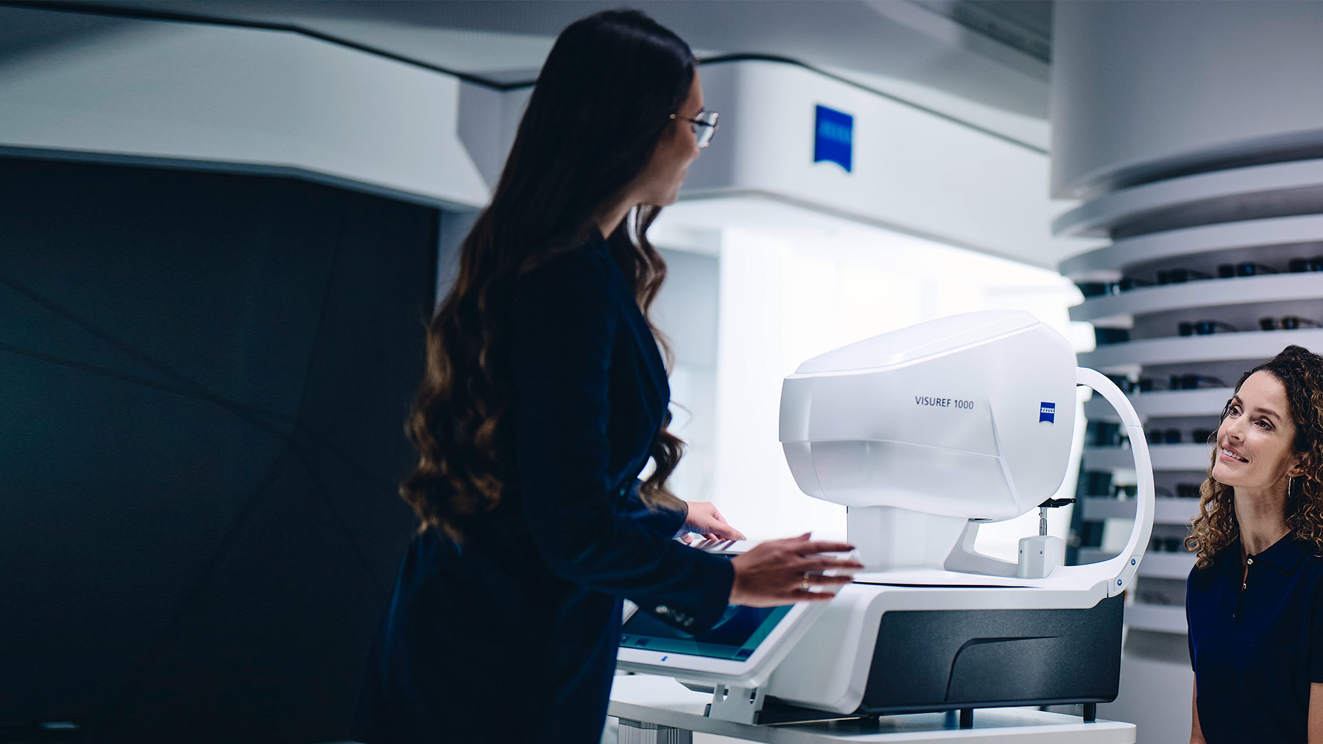 Een vrouw zit voor de ZEISS VISUREF 1000 en kijkt naar een vrouwelijke opticien.