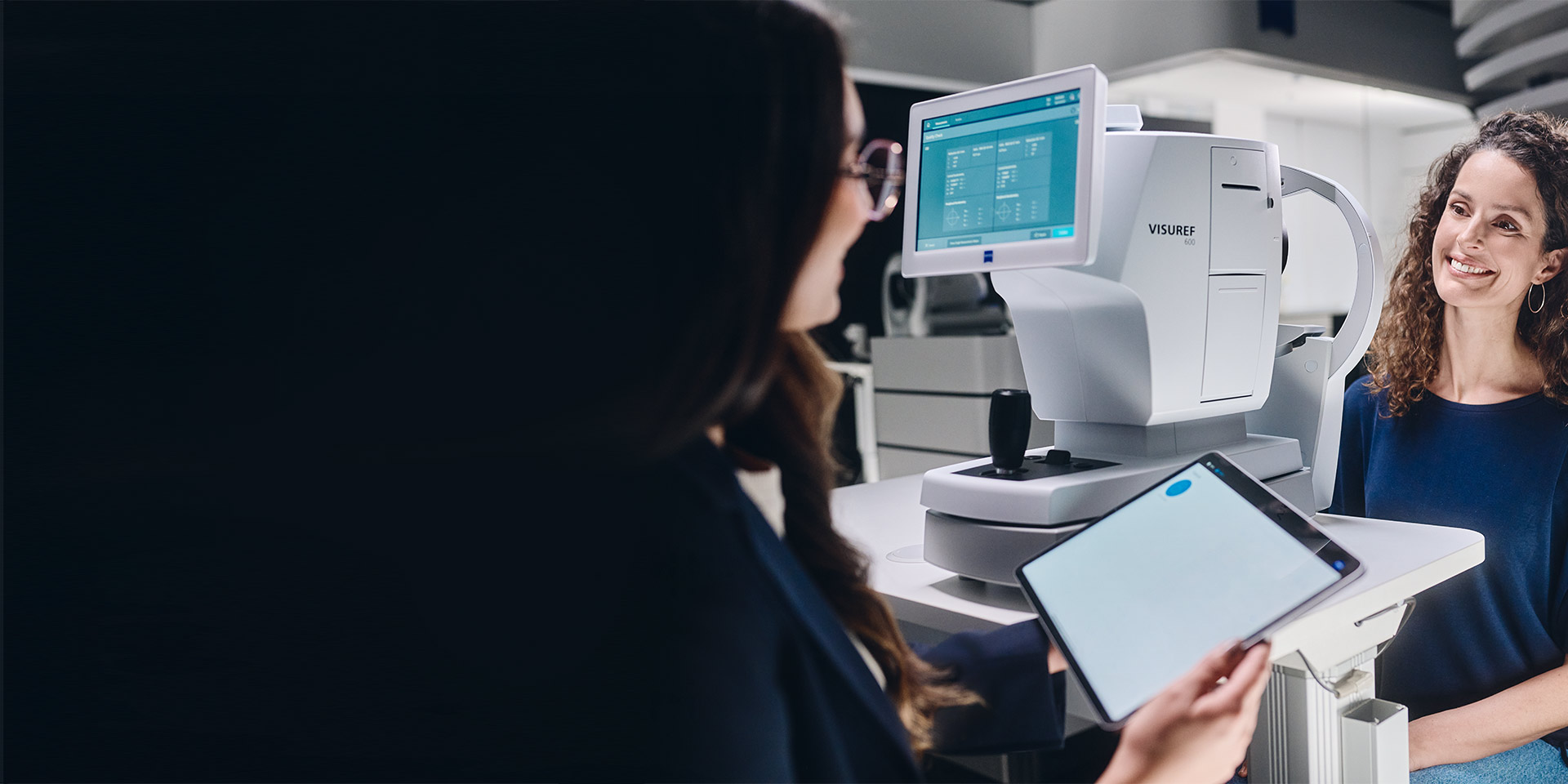 Een vrouwelijke opticien kijkt op haar tablet terwijl ze voor de ZEISS VISUREF 600 zit en met haar klant praat.