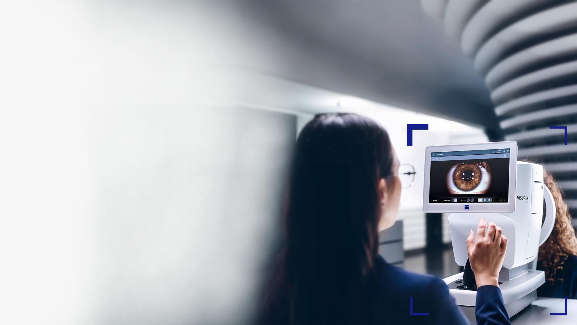 Een vrouwelijke opticien kijkt naar het scherm van de ZEISS VISUREF 600 waarop een beeld van een oog te zien is.