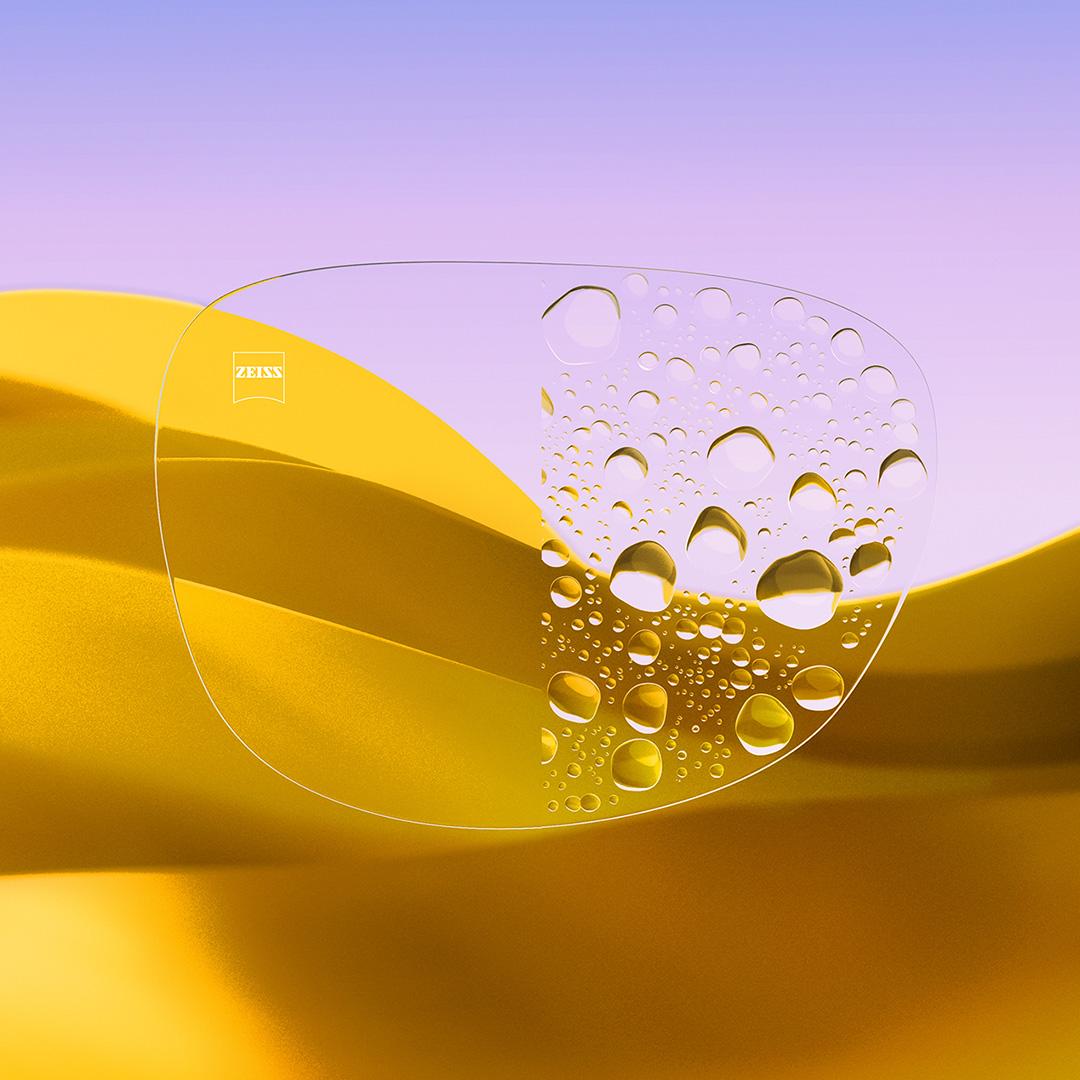 Een afbeelding met een gedeeld brillenglas waarvan de ene helft helder en schoon is dankzij ZEISS DuraVision Gold UV, terwijl de andere helft bedekt is met waterdruppels.
