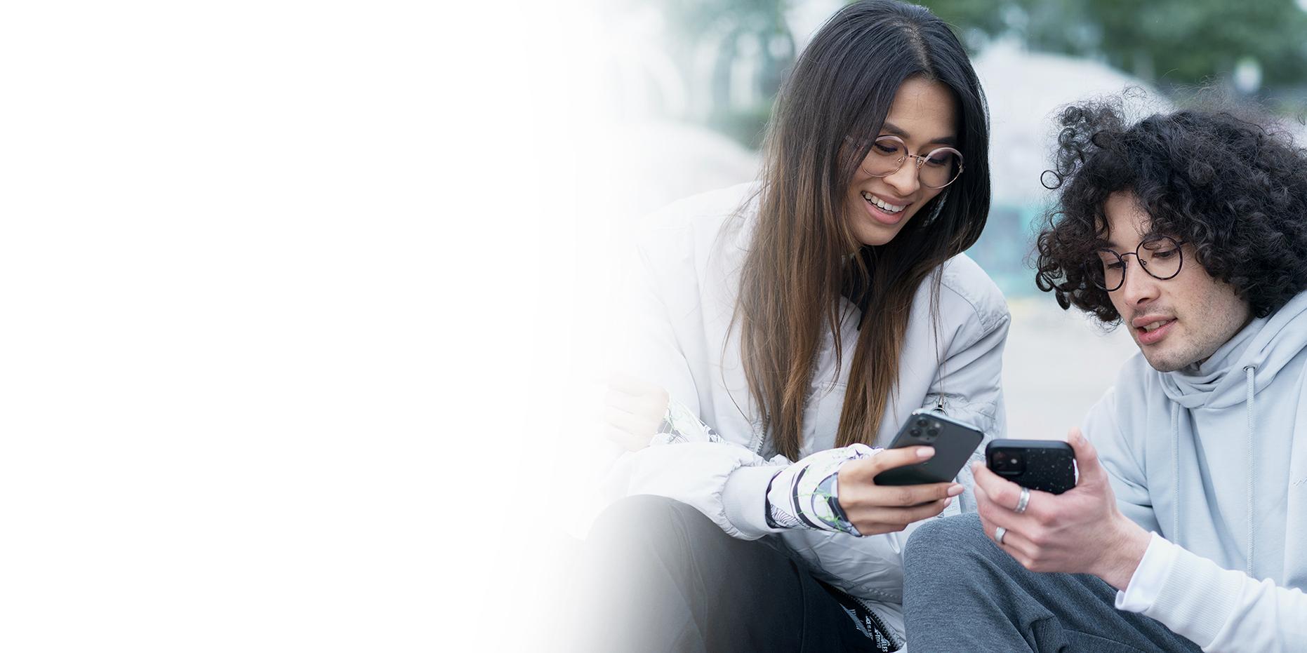 Twee jonge mensen, een vrouw met lang bruin haar die glimlacht en een jongen met krullend zwart haar die een ZEISS SmartLife bril draagt, kijken naar hun telefoon. 