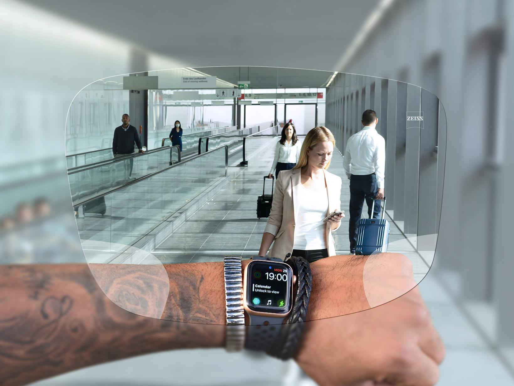Persoon kijkt naar zijn horloge door ZEISS Digital SmartLife brillenglazen. Op de achtergrond zie je een typische luchthavensituatie met mensen die op roltrappen lopen en hun koffers duwen. 