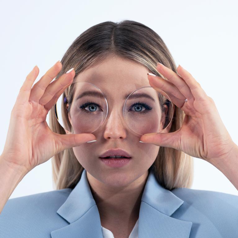 Een jonge blonde vrouw houdt brillenglazen voor haar ogen om het kleine-ogen-effect te laten zien dat veroorzaakt wordt door een dikke min-glazen.