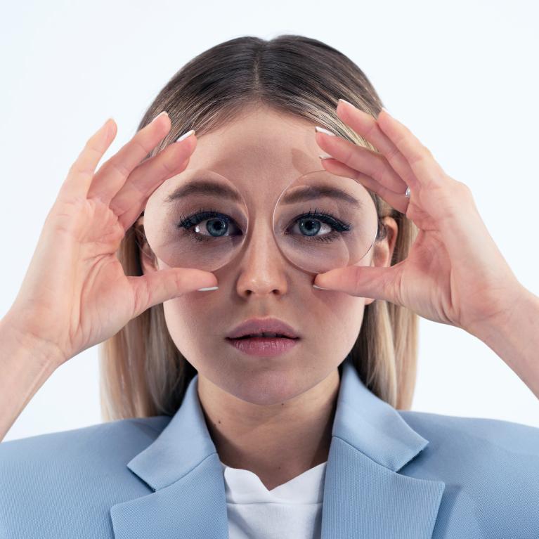 Een jonge blonde vrouw houdt brillenglazen voor haar ogen om het fisheye-effect te laten zien dat veroorzaakt wordt door een dikke bril.