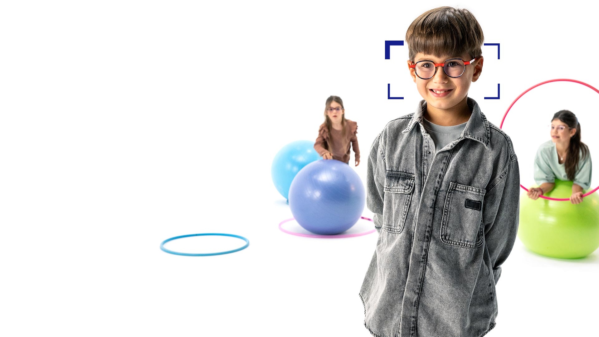 Bruinharige jongen met ronde bril met ZEISS MyoCare glazen staat op de voorgrond en glimlacht naar de camera. Op de achtergrond spelen twee meisjes met ZEISS MyoCare brillenglazen met hoepels en gymnastiekballen.